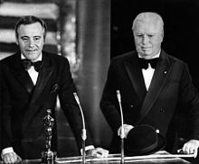 Photographie de Chaplin au visage bouffi et en costume de soirée derrière un pupitre où se trouve une statuette et deux micros