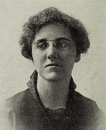 A white woman with short wavy hair, wearing eyeglasses and a dark blouse with a high lace-trimmed collar