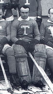 Photo de Harry Hap Holmes en tenue de hockeyeur assis sur un bac au milieu de ses coéquipiers.