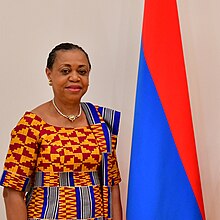 An African woman in traditional dress stands beside a red and blue flag