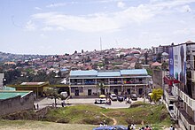 Blick von einer Anhöhe hinunter auf eine belebte Straße mit einem zweistöckigen Gebäude und einer Tankstelle. Im Hintergrund ist ein mit Häusern bebauter Hügel der Stadt.
