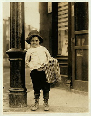 Eight year old newsboy in a black and white photograph with a bundle of newspapers under his arm