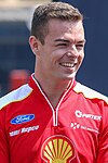 A man in his mid-twenties wearing a red zip T-shirt with a white colour scheme on the shoulders with sponsors logos and looking away from the camera with a smile