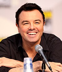 A man with short black hair and a black shirt in front of a microphone. His arms are crossed, and he is laughing.