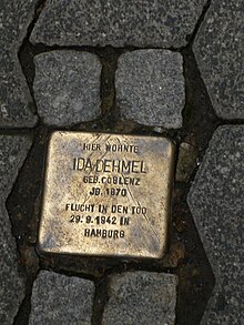 Stolperstein en la casa de Ida Dehmel. Se ve el suelo empedrado y en el centro de la imagen, la piedra de tropiezo en color bronce con la leyenda: «Hier wohnte IDA DEHMEL» y más detalles que ya están borrosos.
