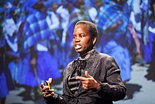 Photograph of an African woman in a grey brocaded suit standing in front of a backdrop depicting a group of African women in blue dresses, while giving a speech. She is wearing a geometrically patterned beaded neck collar, has a yellow device in her right hand, and is pointing with her left index finger to the left of the stage.