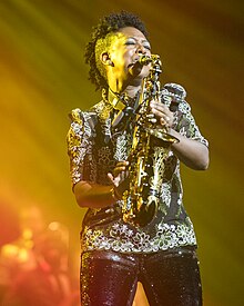 A woman playing the saxophone with eyes closed. Lit from the side with yellow and lit overall in white, she is wearing a black shirt with floral patterns in white and green embroidery and glittery black trousers with Natural hair in a wide, short mohawk, shaved on either side. Behind her, some instruments are out of focus.