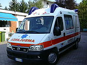 An Italian ambulance, displaying flashing lights in the roof, above the driver's door, and in the front grill; a siren speaker on its roof, striping; the Star of Life; and the reversed "Ambulance" sign on its hood (see text)