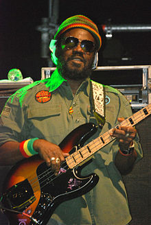 Zdjęcie z 2010 roku. Aston Barrett zwrócony jest w stronę fotografa i gra na czterostrunowej gitarze basowej. Mężczyzna ubrany jest w okulary przeciwsłoneczne, zieloną koszulę typu wojskowego oraz ozdoby w kolorystyce czerwono-zielono-żółtej.