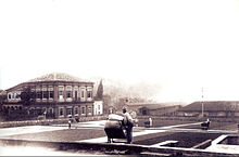 A photograph showing workers spreading or gathering coffee beans drying on a large paved plaza with an elegant, two-story neoclassical building on the left and warehouses and other plantation buildings in the background