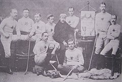 Группа мужчин в форме своей команды, некоторые стоят и некоторые сидят, каждый с хоккейной клюшкой, в студии. На мольберте - баннер чемпионата Оттавы. На столе трофей.