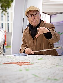 Mann mit Brille, Mütze und braunem Mantel steht vor Kölner Stadtmodell und deutet auf einzelne hervorgehobene Regionen.