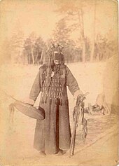 Ancienne photographie sépia, sur fond de paysage estompé, d'un chaman toungouse, debout, portant sa tunique cérémonielle et un casque fort orné dont l'avant de prolonge par un voile qui masque le visage, et tenant de la main son bouclier et de la gauche un bâton sacré.