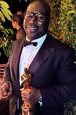 A black male wearing a tuxedo is seen holding an Oscar.