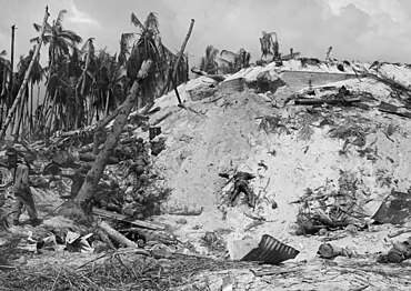 alt = A black-and-white photograph of Tarawa Island. A huge heap of sand can be seen, with numerous dead-bodies of Japanese soldiers.