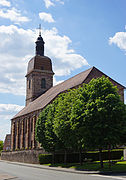 Vue arrière d'une grande église en grès rose surmontée d'un clocher comtois et d'un carillon.