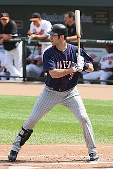 A man in a left-handed batting stance wearing pinstriped gray pants, a black shinguard on his right leg, a dark blue baseball jersey, and a dark-colored batting helmet.