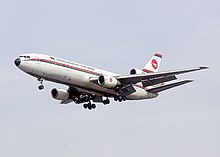 A white plane with red and green cheat lines across its fuselage with landing gears down shown against a blue sky