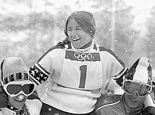 Photographie en noir et blanc montrant une skieuse avec le dossard no 1 portée en triomphe par deux autres personnes.