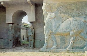 View of a grey stone wall and archway, with the statues of three lamassu (protective deities with wings, the head of a human and the body of a lion or bull).