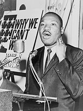 Черно-белая фотография Мартина Лютера Кинга-младшего, выступающего на подиуме, на фоне увеличенной картонной обложки его книги «Почему мы не можем ждать».