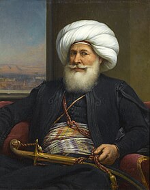 A man with a full white beard and long trained mustache faces the viewer. He wears a white turban and back robe. High on his waist is a gold sash decorated with purple and orange stripes. His left hand holds a cord that goes across his chest, and is connected to a sheathed sword in front of him.