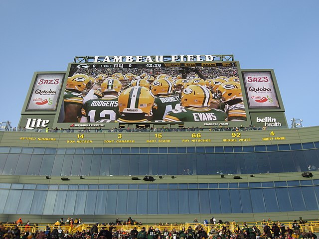 A photo of the stands at Lambeau Field, with a row of the Packers' retired numbers and player names located just above the suite seats. A large video screen is located above the numbers.