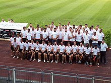 Une quarantaine de joueurs vêtus de t-shirts blancs posent sur une estrade pour une photo de groupe.