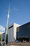 An angled view of a 3 story glass building, bisected by a wide, blank stone wall. There is a slender steel sculpture on the left.