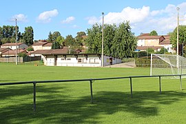 Une terrain de football en herbe derrière une main courante avec une cage de football et un bâtiment dans le fond faisant office de vestiaires.