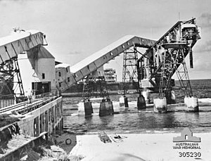 Black and white photograph of an industrial facility that juts out over a body of water. The facility appears damaged as large sections of it are sloping into the water.