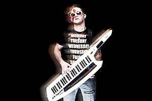 Stylized image of Yinon Yahel with black background, wearing a black t-shirt with white lettering, ripped blue jeans, and white plastic-frame sunglasses, playing a portable keyboard strapped over his shoulder