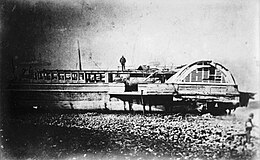 The stern of Princess Alice beached on the shores of the Thames. The paddles wheels are clearly visible, and a man stands on top of the wreckage.