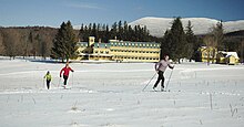 This image contains 3 skiers skiing through a field in front of the yellow buildings at Rikert Outdoor Center