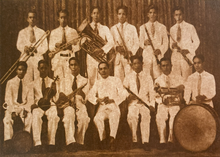 A black and white image of musicians while holding their instruments. The first row in front is seated and the second row at the back is standing.