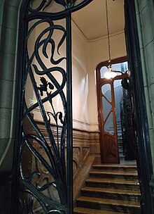 Vista nocturna de la entrada abierta desde la vereda, se pueden ver los marmoles de las escaleras y muros internos del hall, la puerta de madera con diseño orgánico y al fondo el ascensor.