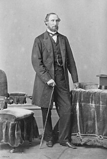 Fair-skinned man with dark hair and beard, standing in a formal portrait pose, wearing mid-Victorian clothes and holding a cane
