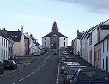 A wide street leads up a slope with parked cares and stone houses painted in whites and yellows on either side. At the end of the street there is a grey and white building with a short spire.