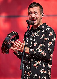 A Caucasian male with brown hair, wearing a floral kimono and singing into a microphone while playing the tambourine.