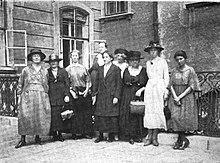Neun Frauen in Kleidern vor einem schmiedeeisernen Zaun stehend, hinter dem sich ein Gebäude mit zwei offenen Fenstern befindet