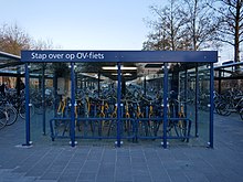 Farbfoto. Frontansicht Fahrradstation. Blau lackierte Stahlkonstruktion mit Glaswänden. Beleuchtet. Beschriftet mit „Stap over op OV-fiets“.