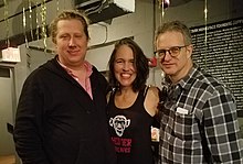 Three people stand in a gray lobby area; on the left, a man in a black sweater and pink button down shirt, to his right, a woman in a black tank top that reads "THEATER OF THE APES", and to her right, a man with glasses in a black and white plaid shirt.
