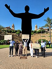 A group of protestors holding signs in front of a modern statue. The statue has been blocked out of the picture it is still under copyright.