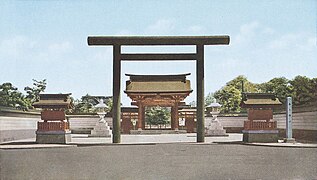 Photo de l'entrée d'un sanctuaire, avec un torii immense à l'entrée, et des lanternes de pierre de chaque côté. Au fond de l'image, on devine le bâtiment principal au bout d'une longue allée rectiligne.