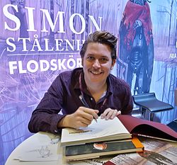 Simon Stålenhag wearing a dark purple patterned shirt with lanyard, sitting at a table in front of a promotional poster of his art, appearing to autograph a book and grinning directly at camera