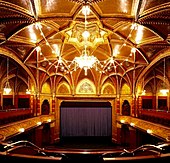 Photographe d'une grande salle comportant une scène avec un rideau foncé, des boiseries et un chandelier au plafond.