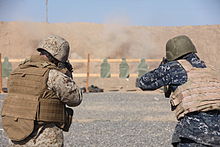 Морской пехотинец (слева) и матрос (справа), одетые в боевое снаряжение, ведут огонь по цели на стрельбище в пустыне.