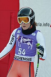 Photo d'une skieuse dans une aire d'arrivée portant casque noir et lunettes de ski, numéro 54 sur chasuble violette décorée d'une vache violette de la marque Milka, en combinaison moulante de ski blanche et orangé