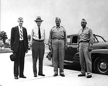 Four men stand in front of a car. The two on the left are wearing suits, the two on the right wear army uniforms with garrison caps and ties tucked in.