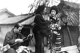 Une femme en kimono sombre (Mitsuko Yoshikawa) arrange les cheveux d'une jeune femme (Kinuyo Tanaka), elle aussi en kimino. Un homme assi (à gauche) répare un shamisen dont le manche est brisé (image en noir et blanc).